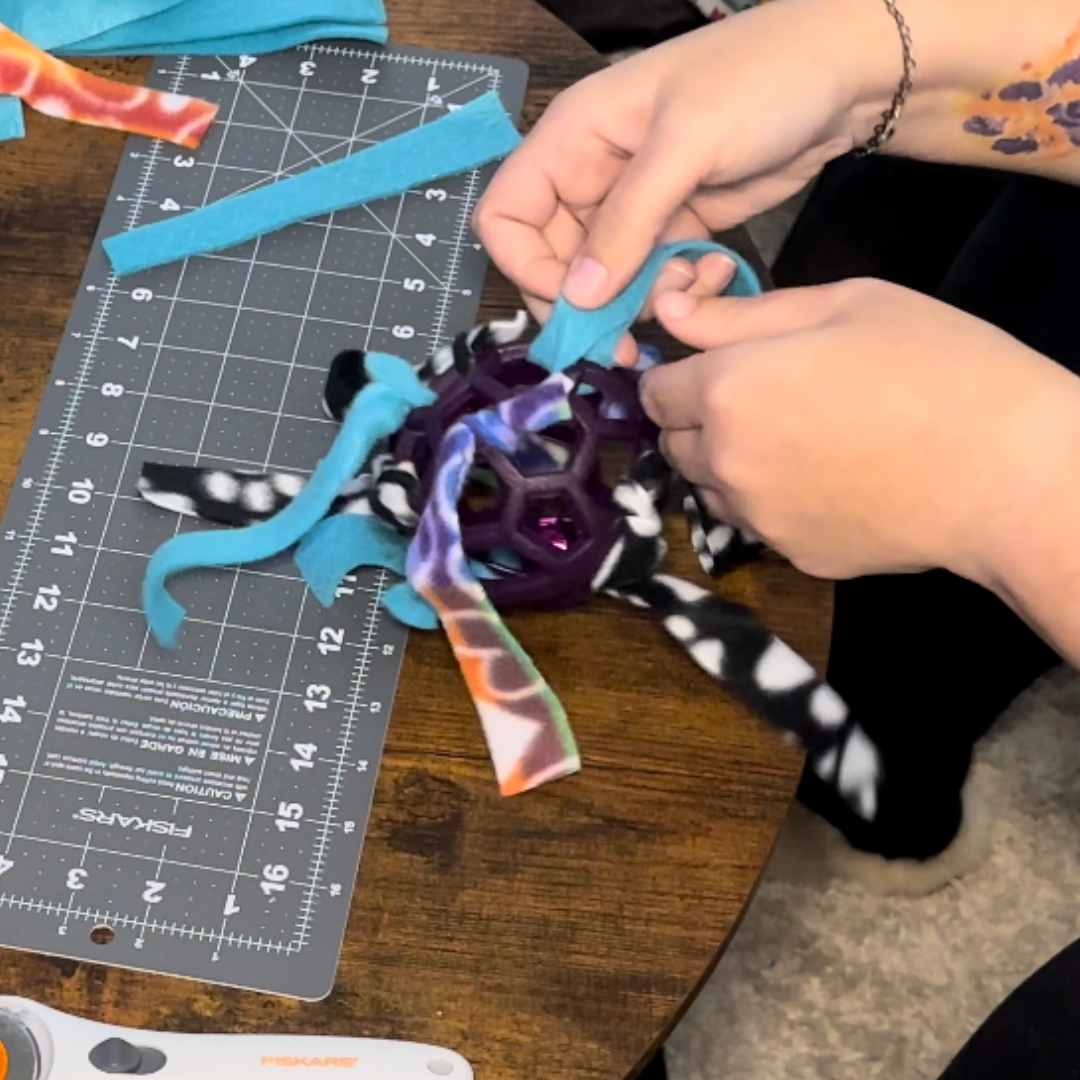A person is attaching fleece strips to a hex ball dog toy on a cutting mat, with scissors nearby for crafting.