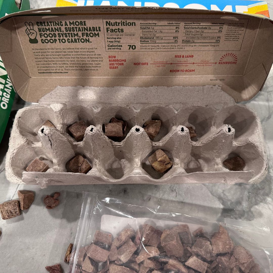 An empty egg carton with dog treats placed in each compartment, alongside a bag of additional treats on a countertop.